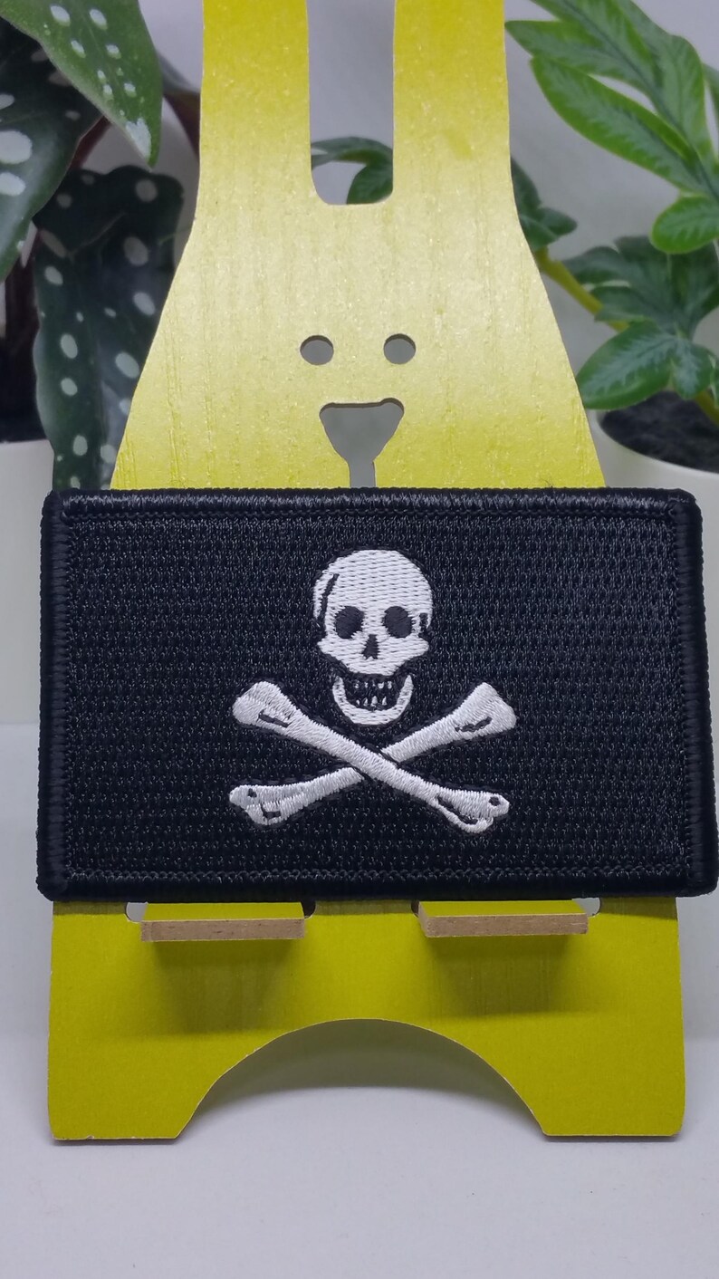 May include: A black embroidered patch with a white skull and crossbones design. The patch is displayed on a yellow stand.