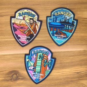 Pode incluir: Três patches bordados em forma de escudo, cada um com a palavra "RANGER" no topo. Os patches apresentam diferentes designs: uma figura com um sabre de luz, uma paisagem futurista e uma estrutura geométrica. As palavras "NATIONAL PARK SERVICE" estão na parte inferior.