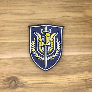 May include: A navy blue embroidered patch with a shield design. The shield features a yellow knight's helmet, a sword, and a crown. The design is framed by yellow laurel branches and white accents. The patch has a black border.