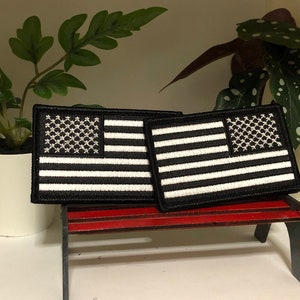 May include: Two black and white embroidered American flag patches on a small red wooden bench. The patches are sewn onto black fabric.