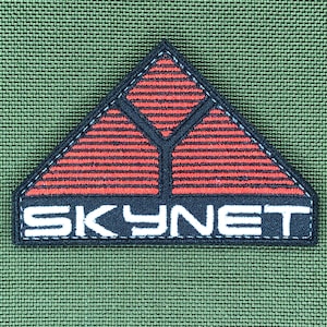 Peut inclure: Écusson brodé avec le mot "SKYNET" en lettres blanches. Le patch présente un motif géométrique rouge et noir à l'intérieur d'un triangle noir, sur un fond vert texturé.