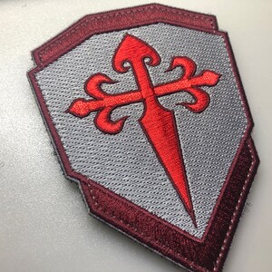 May include: A red embroidered cross with a pointed tip, on a gray shield with a maroon border. The cross is a symbol of the Knights Templar.