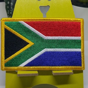 Puede incluir: Un parche de la bandera de Sudáfrica con un borde dorado. La bandera está bordada con los colores rojo, blanco, azul, verde y negro.