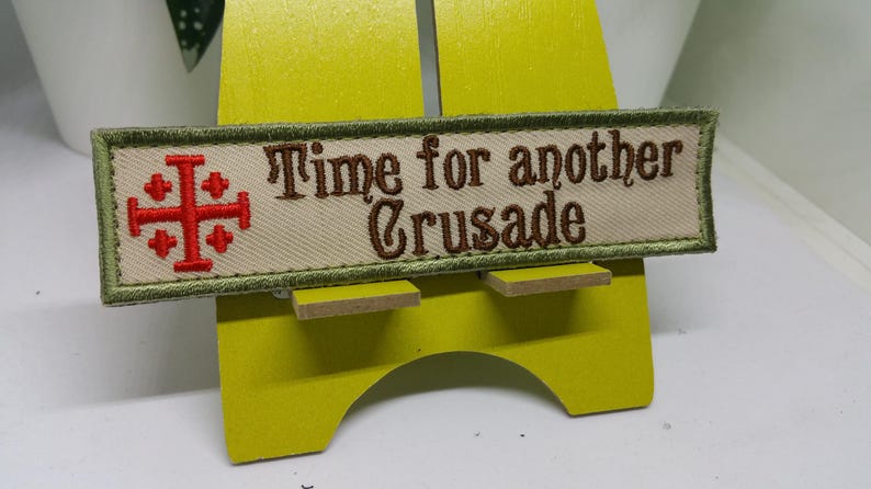 Pu&ograve; includere: Una toppa ricamata verde e beige con una croce rossa e il testo "Time for another Crusade".