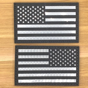 Könnte beinhalten: Zwei schwarz-weiße amerikanische Flaggenaufnäher. Die Aufnäher zeigen das Sternen- und Streifenmuster mit weißen, reflektierenden Streifen und Sternen auf schwarzem Hintergrund. Die Aufnäher sind rechteckig und haben ein strukturiertes Aussehen.