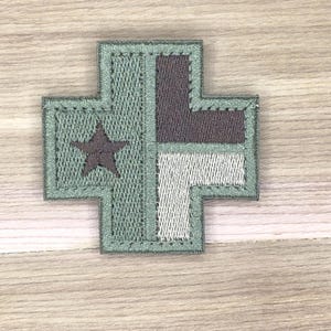 Può includere: Toppa ricamata a forma di croce con una stella. La toppa presenta una combinazione di colori verde oliva, grigio e bianco. La stella è di colore grigio scuro. La toppa è probabilmente per un'uniforme o un capo di abbigliamento.