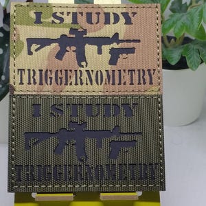 May include: Two green and brown camouflage patches with black stitching. The top patch has a silhouette of a rifle and the text "I STUDY TRIGGERNOMETRY". The bottom patch has the same design but in a darker green color.