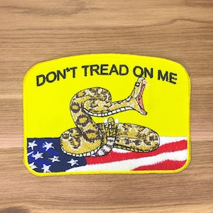 Può includere: Una toppa gialla con un serpente a sonagli arrotolato e la scritta "DON'T TREAD ON ME". Il serpente è posto su uno sfondo della bandiera americana. La toppa è rettangolare con angoli arrotondati. Il testo è nero.