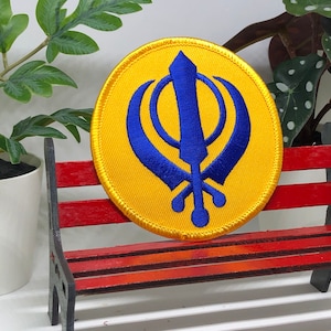 May include: A yellow embroidered patch with a blue Khanda symbol, a Sikh religious symbol, on a red wooden bench.