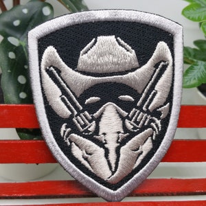 Puede incluir: Parche bordado con forma de escudo, con un sombrero de vaquero, pistolas cruzadas y un diseño de calavera. El parche tiene un fondo negro con costuras grises y blancas, y un borde plateado. El parche está colocado en un banco rojo.