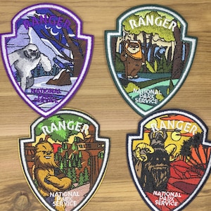 Pode incluir: Quatro patches bordados Ranger do Serviço de Parques Nacionais. Cada patch apresenta um personagem ou cena diferente, incluindo um yeti, um Ewok, Chewbacca e um Wampa. Os patches têm formato de escudo com uma borda branca e as palavras "Ranger" e "National Park Service".