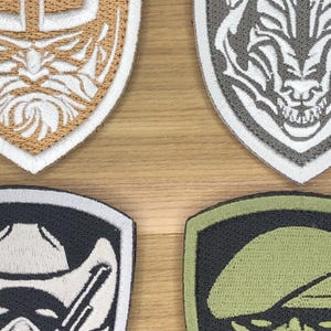 May include: Four embroidered patches in shield shapes. The top left patch is tan and white with a detailed design. The top right patch is grey and white with a skull design. The bottom left patch is black and white. The bottom right patch is green and black.