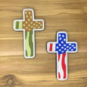 Pode incluir: Dois patches bordados em forma de cruz. Um apresenta um padrão de camuflagem com estrelas douradas, o outro exibe o design da bandeira americana com listras vermelhas e brancas e estrelas azuis. Ambos têm uma borda branca.