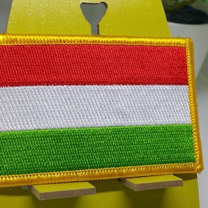May include: A red, white and green embroidered patch with a yellow border. The patch is rectangular in shape and is likely a national flag.