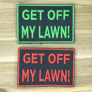 Pode incluir: Duas etiquetas retangulares com o texto "GET OFF MY LAWN!". Uma etiqueta tem texto e borda verdes sobre fundo preto. A outra tem texto e borda vermelhos sobre fundo preto. As etiquetas estão sobre uma superfície de madeira clara.
