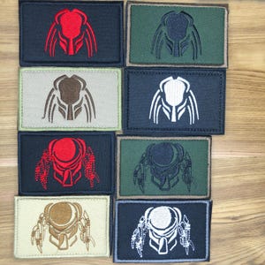 May include: A collection of embroidered patches featuring the Predator's mask design. The patches are rectangular, with various color combinations including red, white, and brown on black, green, and tan backgrounds. The patches are likely for clothing or accessories.