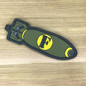 Può includere: Toppa ricamata verde oliva a forma di bomba. La toppa presenta un cerchio giallo con una "F" nera al centro. La toppa ha un bordo nero ed è su una superficie di legno.
