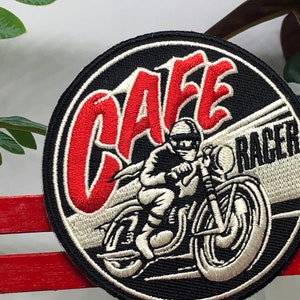 May include: A black and white embroidered patch with red lettering that says "Cafe Racer". The patch features a white motorcycle with a rider in a white helmet.