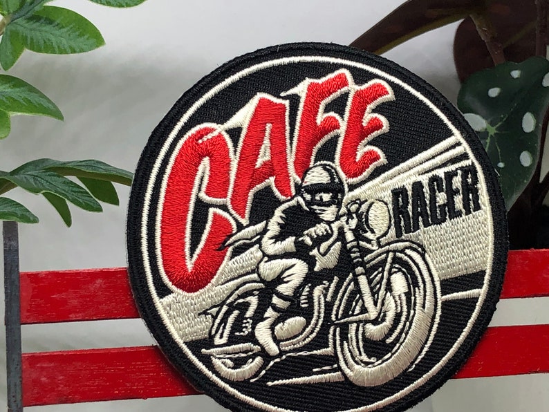 May include: A black and white embroidered patch with red lettering that says "Cafe Racer". The patch features a white motorcycle with a rider in a white helmet.