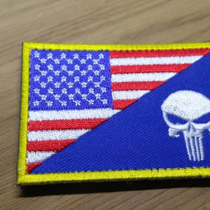 Pode incluir: Um remendo com borda amarela, fundo azul e uma caveira branca. A metade superior do remendo apresenta uma bandeira americana vermelha, branca e azul.