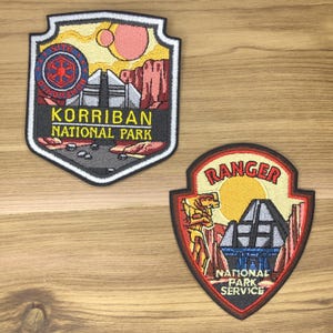 Pode incluir: Dois patches bordados. Um patch diz "KORRIBAN NATIONAL PARK" com um design de escudo, apresentando uma paisagem com formações rochosas vermelhas e uma estrutura geométrica. O segundo patch diz "RANGER NATIONAL PARK SERVICE".