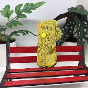 May include: A gold-colored embroidered patch of a hand wearing an infinity gauntlet with gems. The patch is sitting on a red wooden bench.