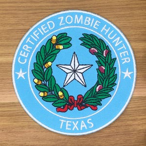 May include: A round patch with a light blue background and white border. The text "CERTIFIED ZOMBIE HUNTER" and "TEXAS" are in white. A white star is in the center, surrounded by a green wreath with yellow and red accents.