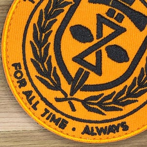 Könnte beinhalten: Runder, orangefarbener Aufnäher mit schwarzem Stickdesign und Text. Das Design umfasst eine zentrale geometrische Form, Olivenzweige und die Worte "FOR ALL TIME • ALWAYS" in einem Kreis.