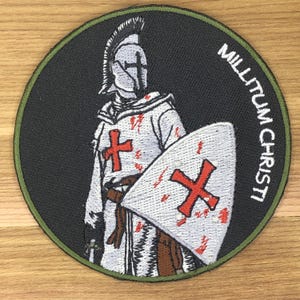 May include: Embroidered patch featuring a knight in white armor with a red cross on the chest and shield. The patch has the words "MILLITUM CHRISTI" in white text on a black background, with a green border.
