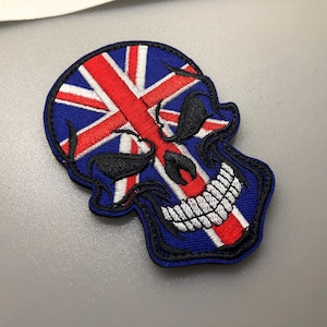 Puede incluir: Parche bordado de una calavera con un diseño de bandera Union Jack. La calavera es azul con dientes blancos y ojos negros. La bandera es roja, blanca y azul.