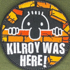 Op de afbeelding: Ronde patch met de tekst "KILROY WAS HERE!" in witte letters. Het ontwerp toont een cartoonfiguur die over een bakstenen muur kijkt. De muur is oranje en geel, en de figuur is zwart met witte ogen.