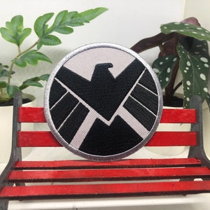 May include: A white and black embroidered patch with a silver outline. The patch features a stylized eagle with wings spread wide, inside a shield shape. The shield is surrounded by a circle.