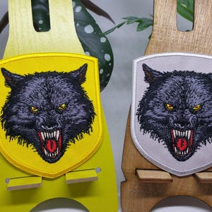 Könnte beinhalten: Zwei gestickte Aufnäher mit einem grimmigen Wolfskopf. Einer hat einen gelben Hintergrund, der andere einen grauen. Beide haben schwarzes Fell, gelbe Augen und rote Mäuler. Die Aufnäher werden auf Holzständern präsentiert.