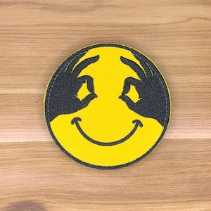 Puede incluir: Un parche redondo amarillo con un diseño de cara sonriente. El diseño incluye dos manos negras haciendo un gesto de 'ok' por encima de los ojos. El parche tiene un borde negro y es probablemente un parche para planchar o coser.