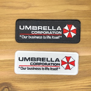 Könnte beinhalten: Zwei rechteckige Aufnäher. Einer ist schwarz mit weißem Text und einem rot-weißen Schirm-Logo. Der andere ist weiß mit schwarzem Text und demselben Logo. Beide Aufnäher tragen den Text "UMBRELLA CORPORATION" und das Zitat "Our business is life itself."