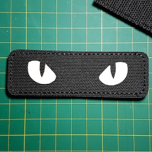Könnte beinhalten: Schwarzer Stoff-Patch mit zwei weißen Augen. Die Augen sind sichelmondförmig.