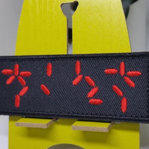 May include: A black fabric patch with red embroidered Japanese characters. The patch is rectangular and features a series of stylized symbols. The background is a bright yellow wooden display.