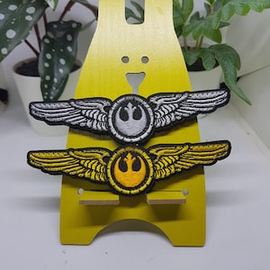 May include: Two embroidered patches featuring a Rebel Alliance symbol and wings. The top patch is white with silver wings and the bottom patch is yellow with gold wings.