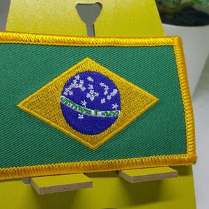 以下が含まれることがあります： 黄色、緑色、青色のブラジルの国旗の刺繍パッチ。パッチは黄色のスタンドに置かれています。