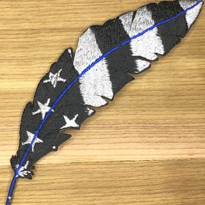 May include: Embroidered feather patch with a black and white striped design, a blue line, and white stars. The patch is on a wooden surface.