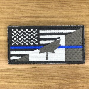 Op de afbeelding: Geborduurde patch met een ontwerp dat de Amerikaanse vlag, een blauwe lijn en een grijs esdoornblad bevat. De patch heeft een zwarte rand en een rechthoekige vorm. Het vlaggedeelte is wit en zwart.