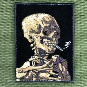 May include: Embroidered patch depicting a skull smoking a cigarette. The skull is detailed with tan and purple thread on a black background. The patch is rectangular with a black border. Suitable for attaching to clothing or bags.