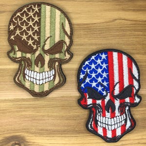May include: Two embroidered skull patches. One features a camouflage pattern with brown and green hues. The other displays the American flag design with red and white stripes and blue stars. Both have white teeth and black outlines.