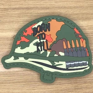 Puede incluir: Un parche de casco militar verde con las palabras "BORN TO KILL" en negro. El diseño del casco presenta un patrón de camuflaje en tonos verdes, marrones y naranjas, con una fila de balas a lo largo del lateral.