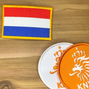 Könnte beinhalten: Gestickte Aufnäher mit der niederländischen Flagge und einem königlichen Emblem. Der rechteckige Flaggenaufnäher zeigt horizontale Streifen in Rot, Weiß und Blau mit einem gelben Rand. Zwei runde Aufnäher zeigen eine Krone und die Buchstaben "KN" in Orange und Weiß.