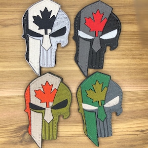 May include: Four embroidered patches in the shape of a Spartan helmet and skull, each with a Canadian maple leaf design. The patches feature a variety of colours, including black, red, grey, green, and tan. The patches are likely for clothing or accessories.