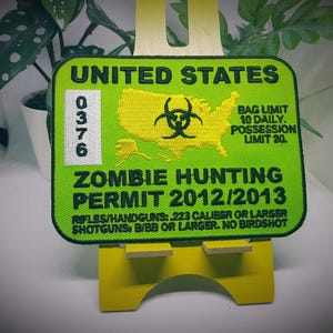 Op de afbeelding: Een groene en gele geborduurde patch met de tekst "UNITED STATES" en "ZOMBIE HUNTING PERMIT 2012/2013". De patch heeft een biohazard-symbool en een kaart van de Verenigde Staten.