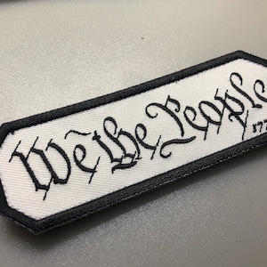 Peut inclure: Un écusson rectangulaire avec un fond blanc et une bordure noire. Les mots "We the People" sont brodés en écriture noire, avec "1776" en dessous. L'écusson a une bordure noire.