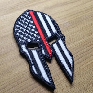 Op de afbeelding: Een geborduurde patch in zwart en wit in de vorm van een Spartaanse helm met een rode streep in het midden. De patch is ontworpen om eruit te zien als de Amerikaanse vlag met een dunne rode lijn.