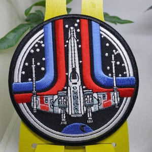 Puede incluir: Parche redondo bordado con un diseño de nave espacial blanca con rayas rojas y azules sobre un fondo negro. El parche tiene un borde blanco con pequeños puntos blancos. El parche se muestra en un soporte amarillo.